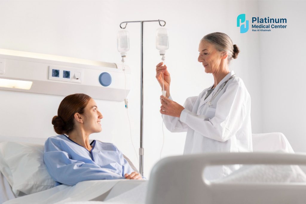 A female patient lying in a hospital bed, looking up and smiling at a healthcare provider in a white lab coat who is adjusting an IV drip bag on a vertical pole.